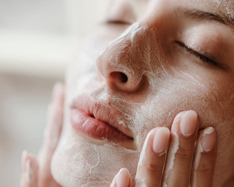 Close-up of a woman's face with a white foaming cleanser or mask applied, being gently wiped with her fingers in a soft, bright skincare setting.