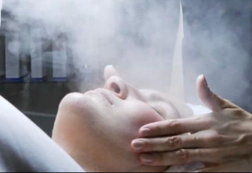 Person receiving a facial treatment with steam and a professional in a dark room.