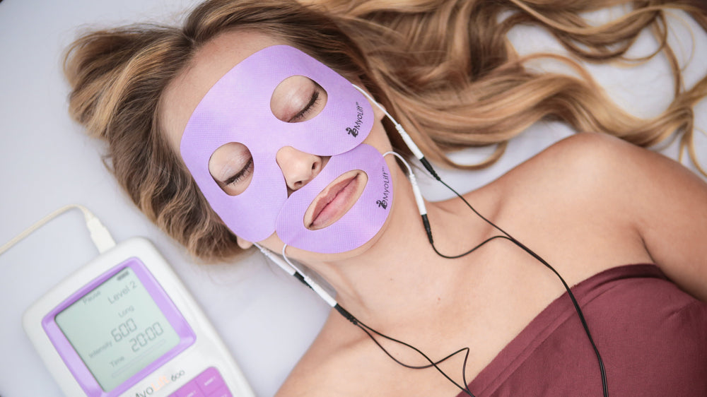 Woman receiving facial treatment with a purple mask and electronic device on a white surface.